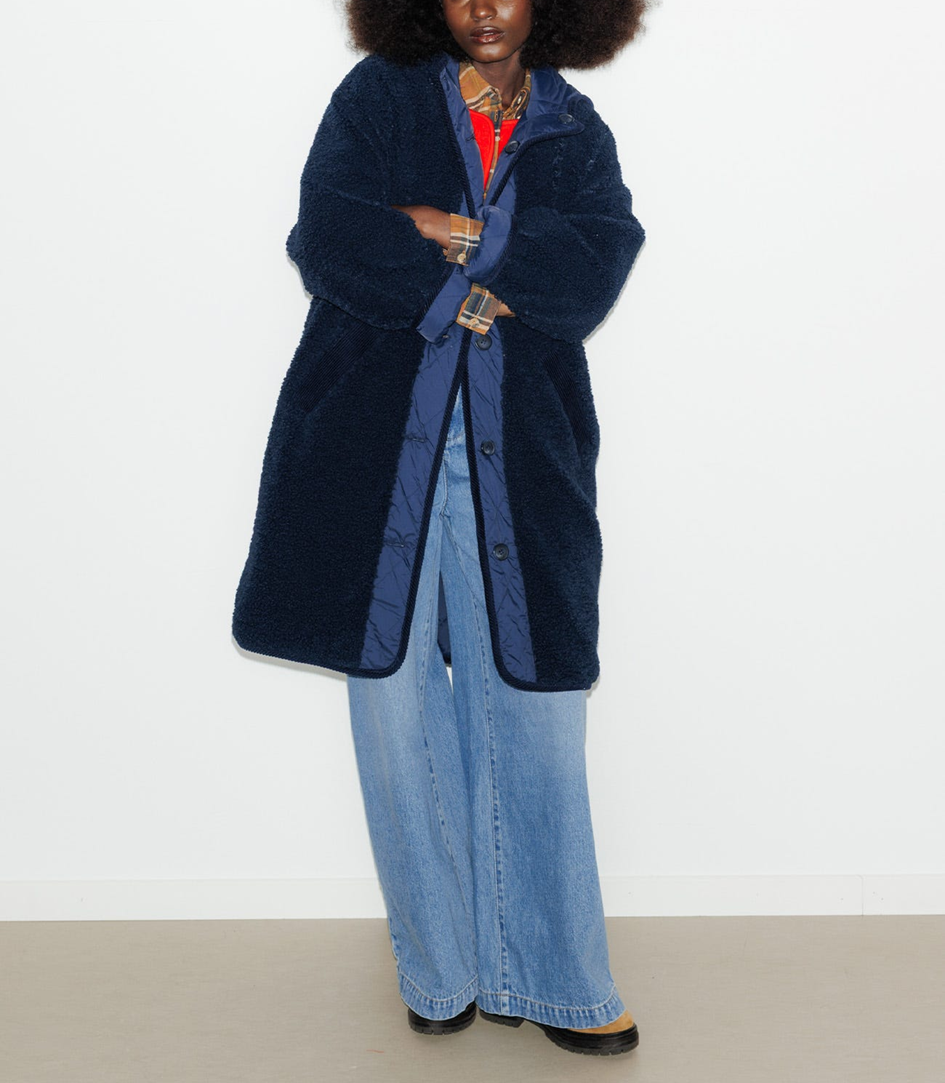 mannequin portant un manteau long reversible avec sa grande capuche. elle est réversible : un côté en moumoute sherpa super doux et un côté matelassé uni classy. Coupe droite