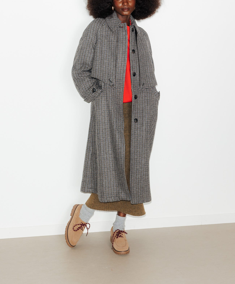 mannequin portant un trench en pied de poule,col pointe à revers, bavolets boutonnés devant et dos, emmanchures raglan, patte boutonnée aux poignets. Coupe longue et évasée. 