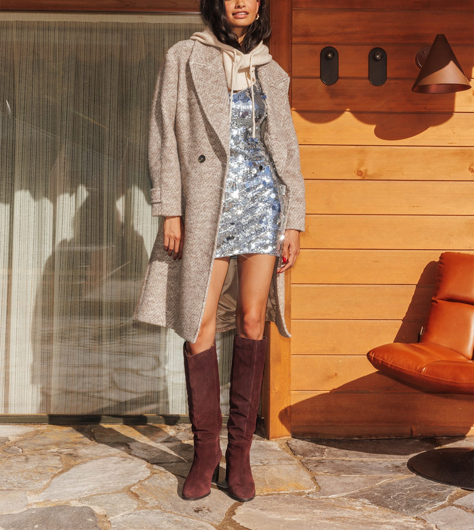 mannequin portant un manteau long en laine beige avec une robe à paillette courte en dessous et des bottes hautes